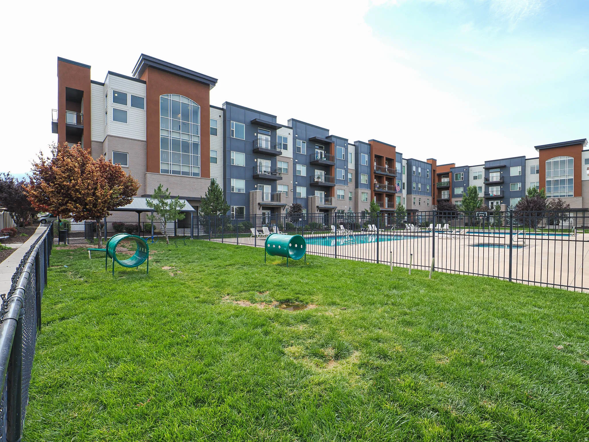 Grassed outdoor amenity space featuring recreational equipment, adjacent to a multi-story building with balconies, and a fenced swimming pool area