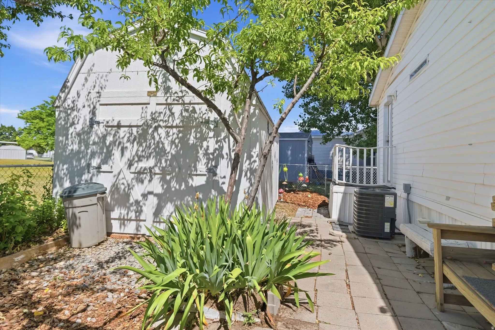 Side yard featuring a white storage shed with double doors, mature tree, and a paved walkway leading to a private deck