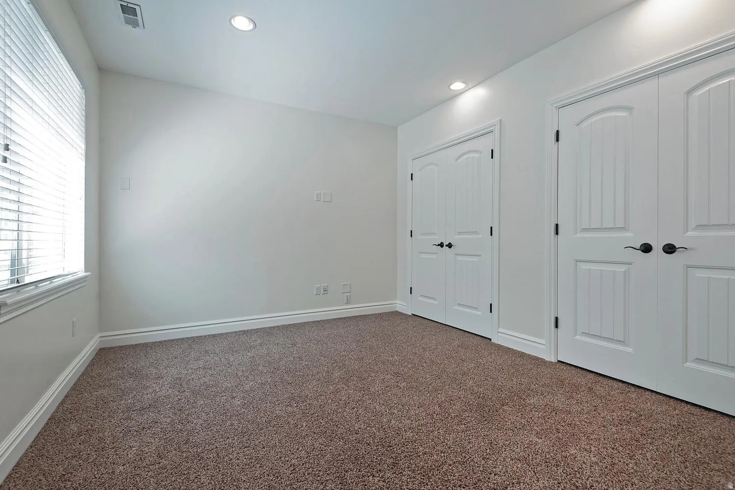 Carpeted room featuring recessed lighting, a window with blinds, two sets of white double bi-fold doors, and white baseboards