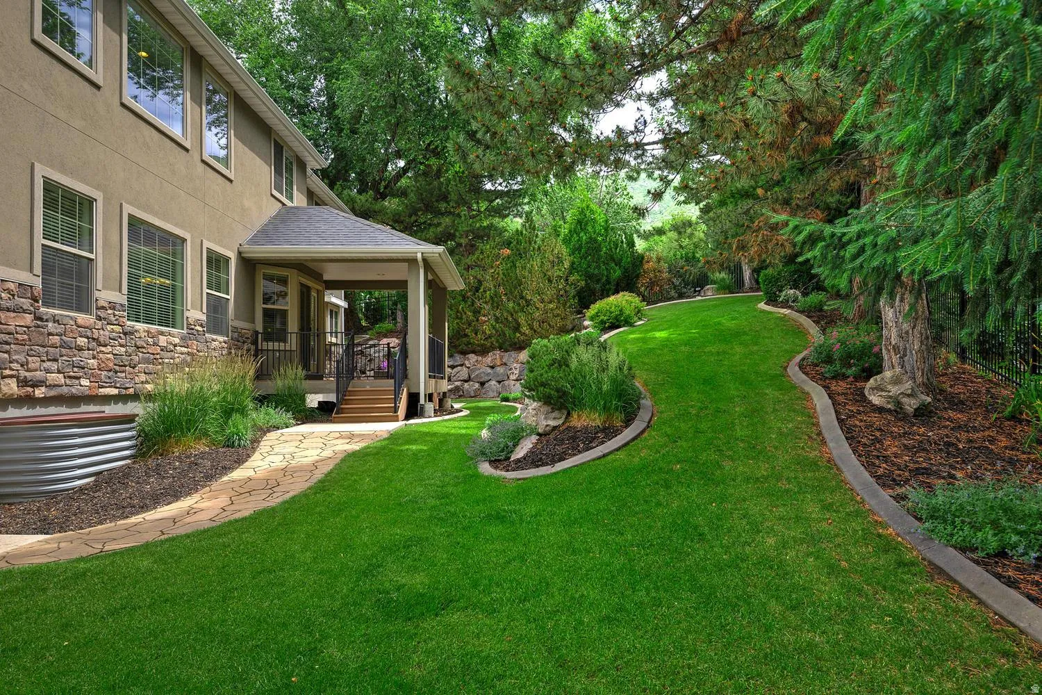 Expansive backyard featuring a winding stone-patterned pathway, landscaped garden beds, a covered porch with steps, and mature trees