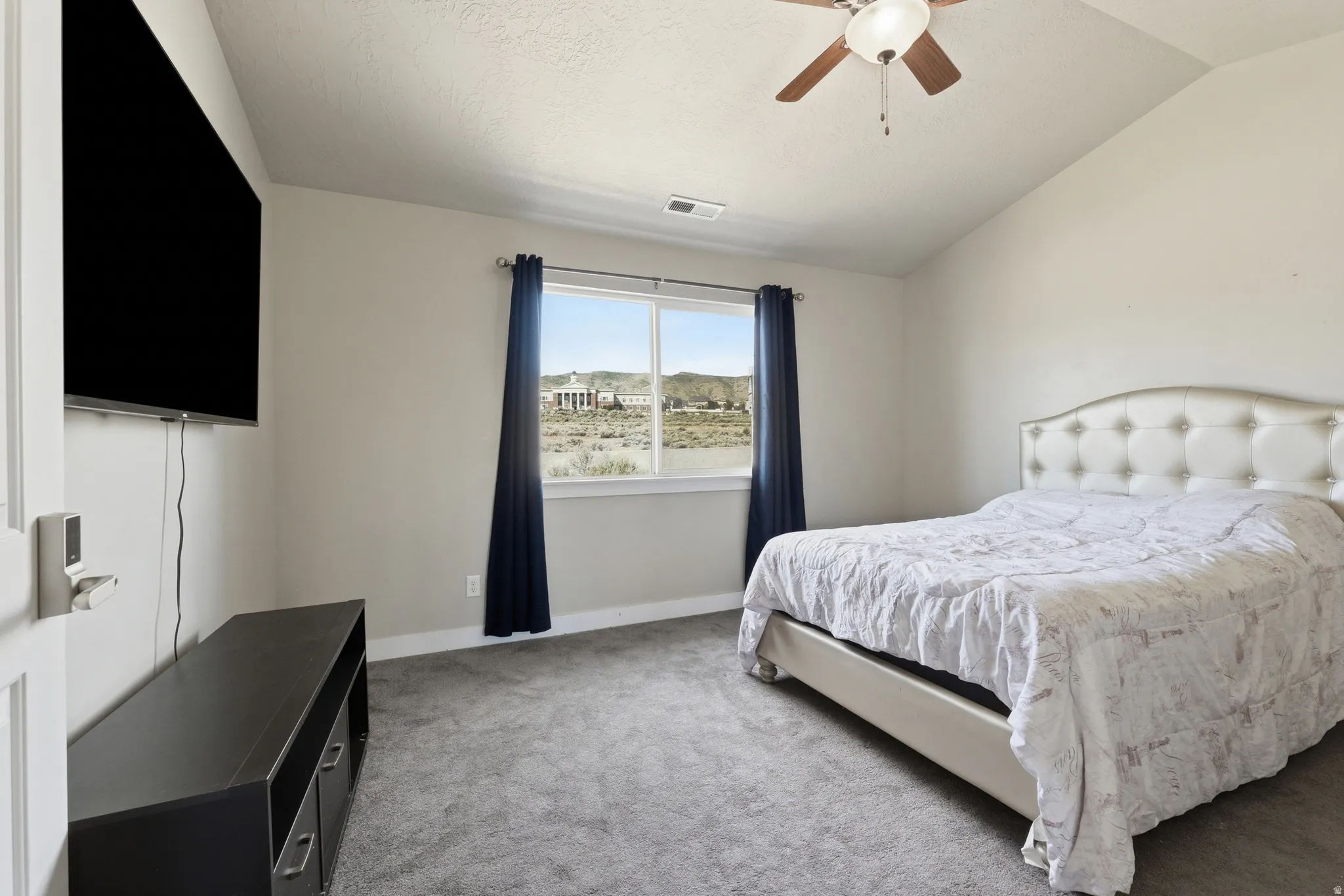 Vaulted ceiling bedroom with a ceiling fan, plush gray carpeting, and a window framing distant landscape