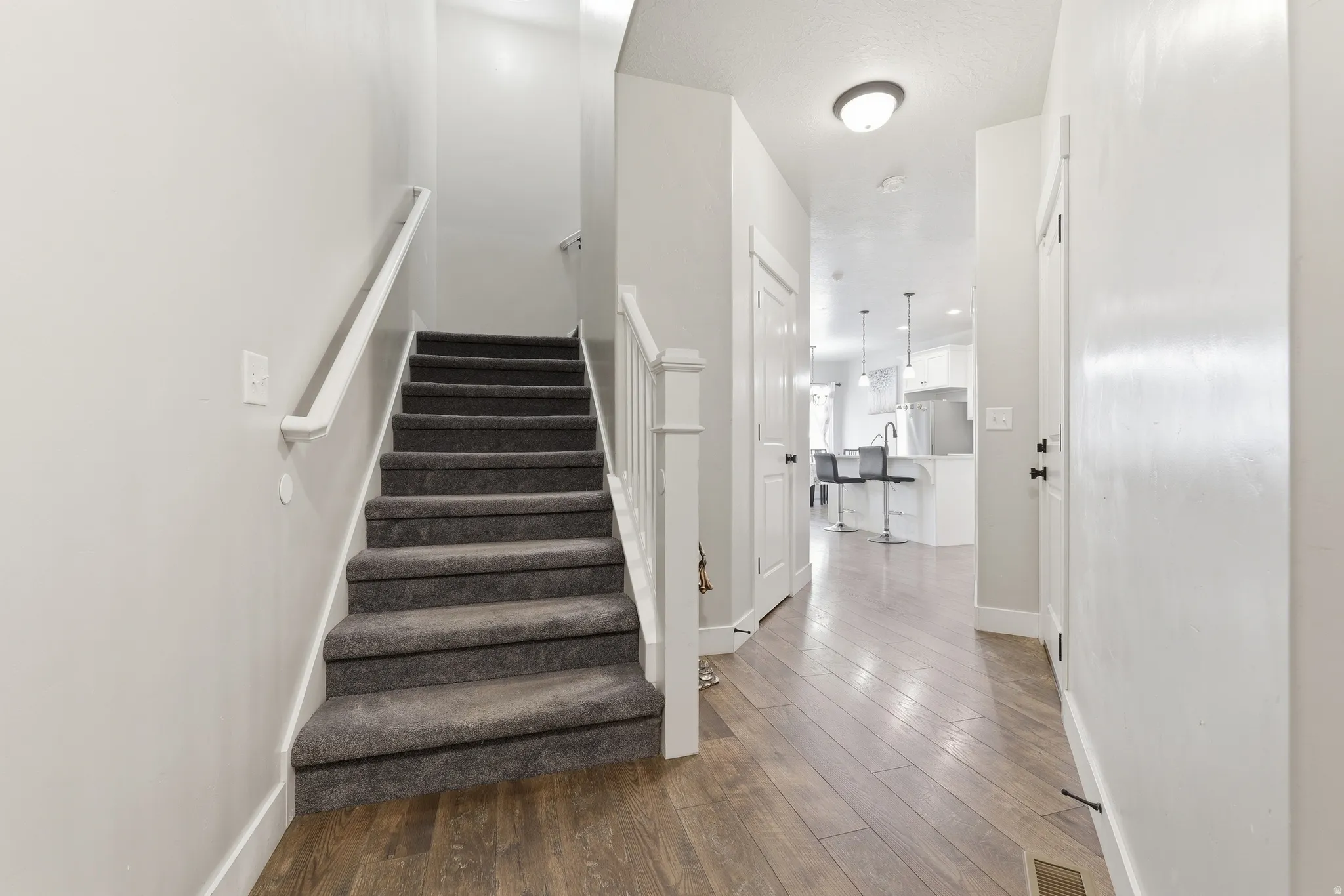 Inviting entryway featuring wood-finish flooring, a carpeted staircase with white railings, and a clear line of sight to a modern kitchen with pendant lighting
