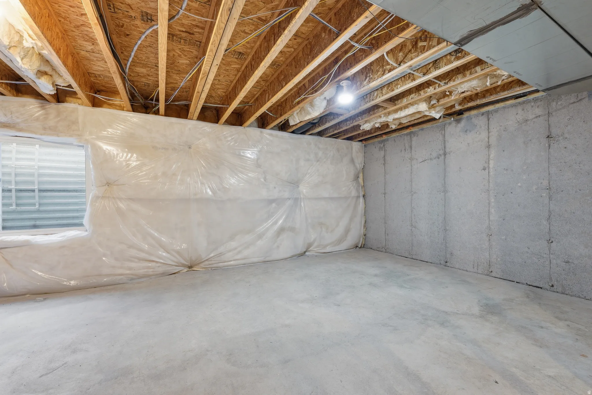 Unfinished basement featuring exposed wood joists, concrete flooring, concrete walls, and a visible window with a gridded well