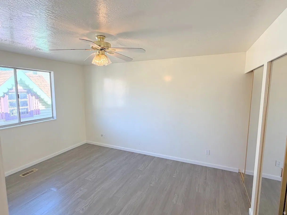 Spacious room featuring light-toned walls, wood-finish flooring, a ceiling fan with integrated lighting, and mirrored sliding closet doors