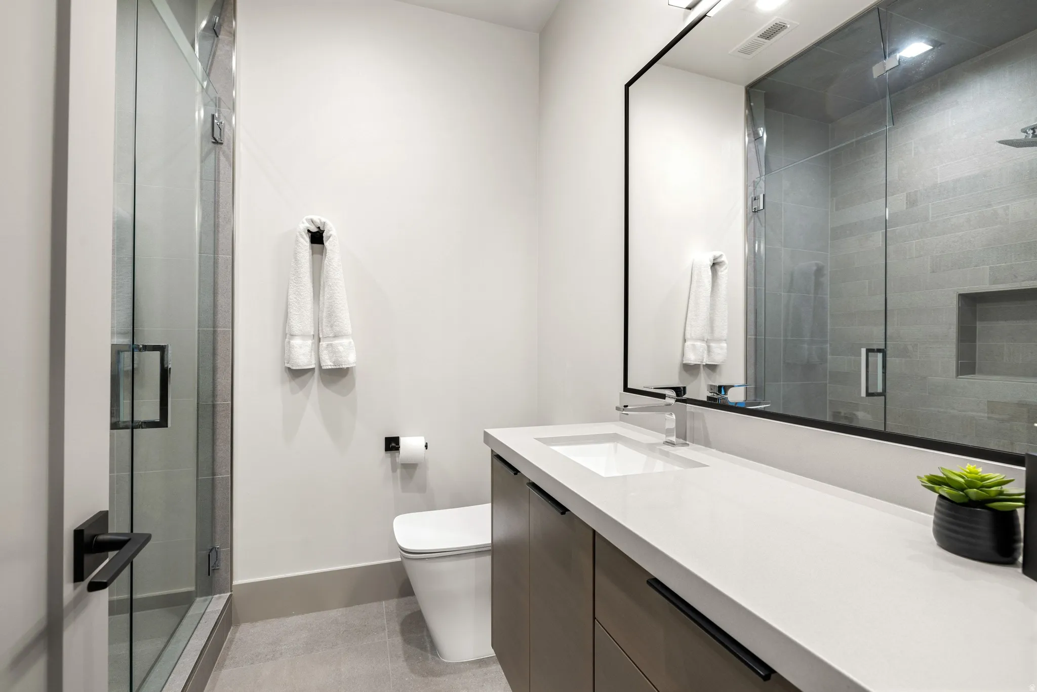 Contemporary bathroom featuring a frameless glass shower enclosure, a sleek vanity with an integrated sink, a rectangular mirror with a black frame, a wall-mounted toilet, and light-toned floor tiling
