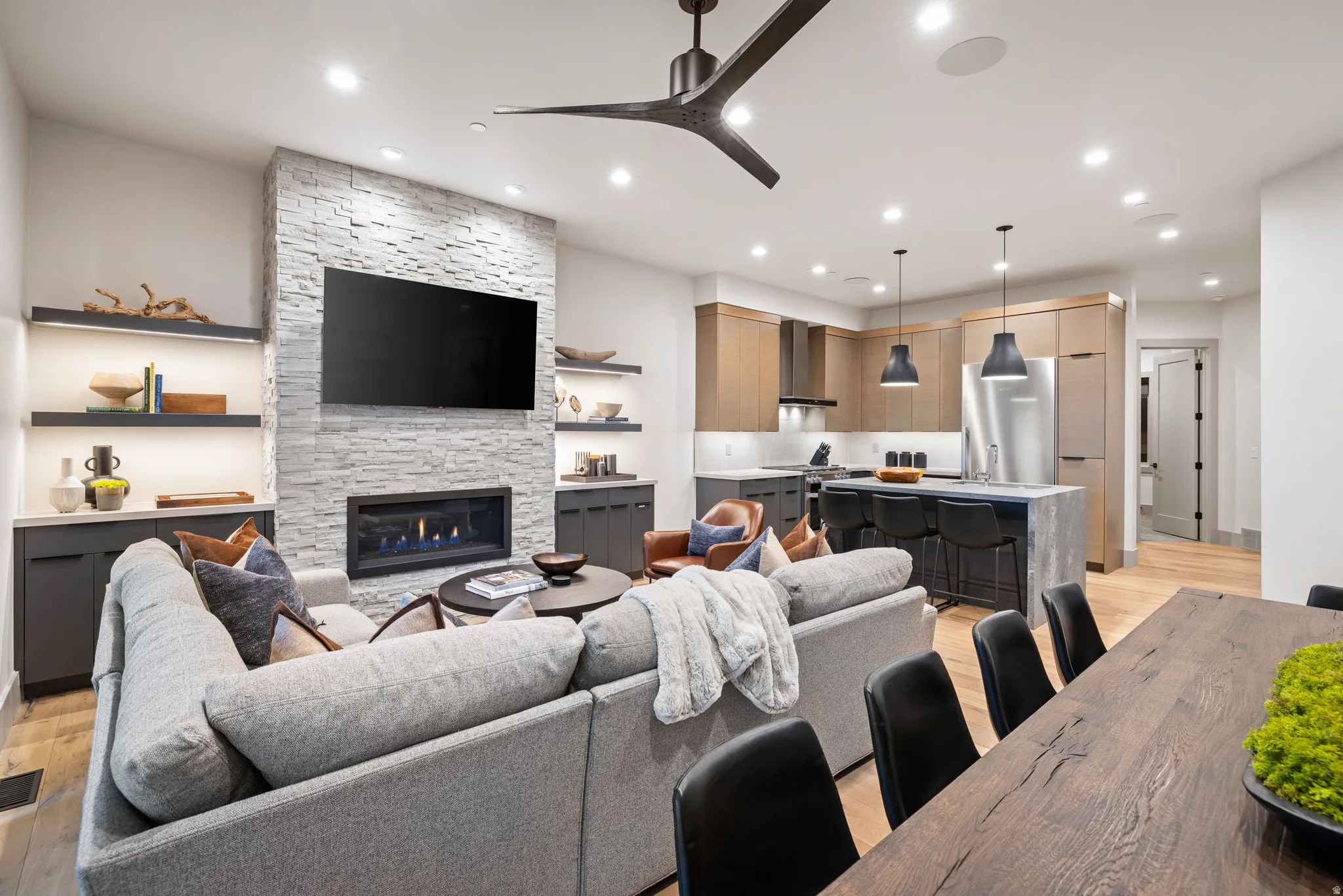 Open-concept living area featuring a stacked stone fireplace with a linear gas insert, built-in shelving with integrated lighting, a modern kitchen with a large island and pendant lighting, stainless steel appliances, and wood-finish flooring