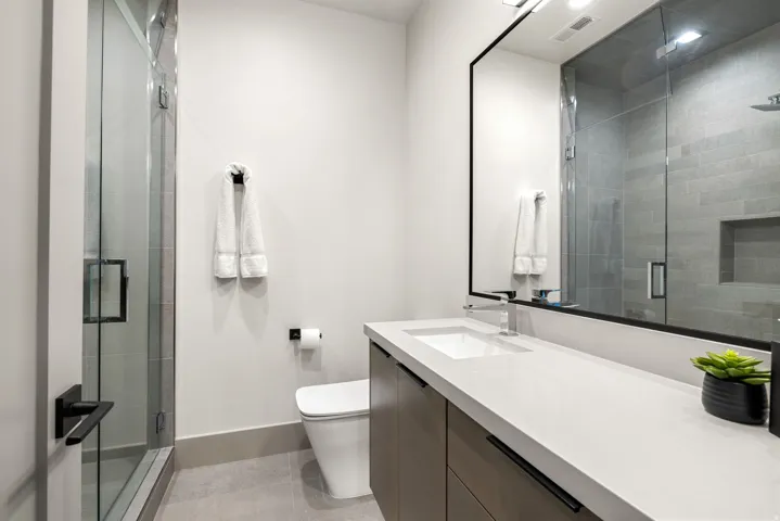 Contemporary bathroom featuring a frameless glass shower enclosure, a sleek vanity with an integrated sink, a rectangular mirror with a black frame, a wall-mounted toilet, and light-toned floor tiling