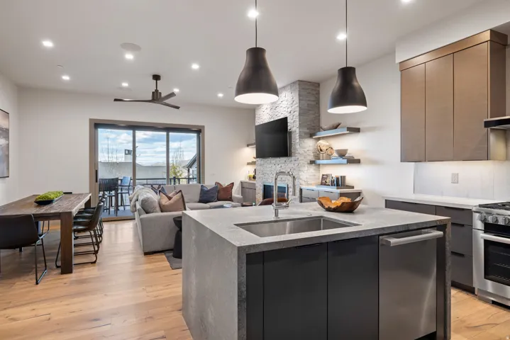 Open-concept living area featuring a kitchen island with integrated sink, wood-finish flooring, a stone-accented fireplace, and sliding glass doors opening to an outdoor space