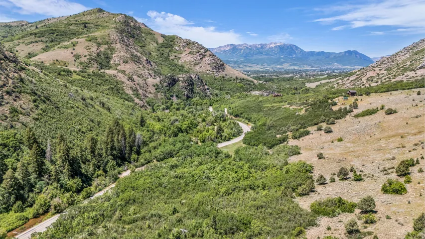 Expansive mountain landscape featuring varied terrain, abundant natural vegetation, a winding road, and distant snow-capped peaks
