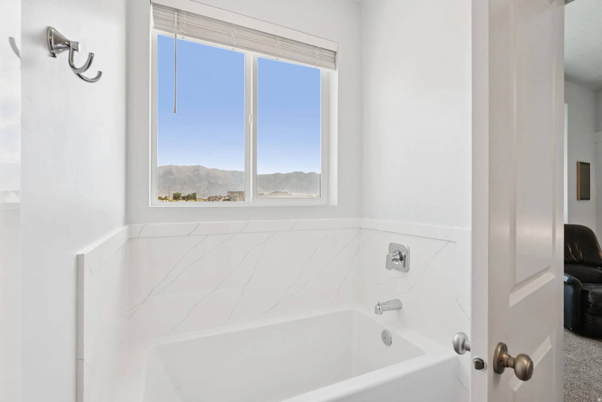White-paneled bathtub featuring a built-in window with mountain scenery, polished chrome fixtures, and white tile surround
