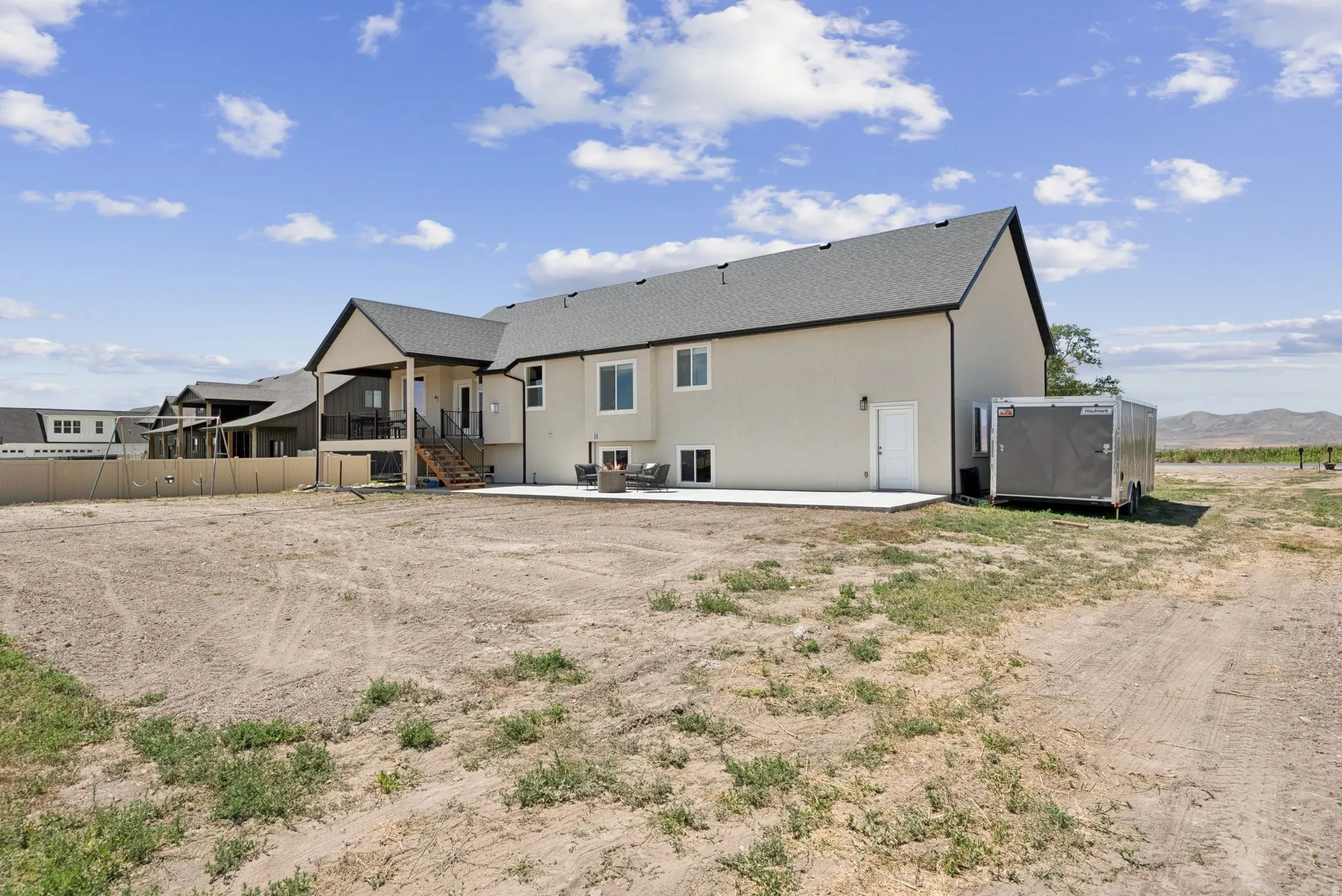 Expansive rear exterior featuring a covered upper deck with black railings, a ground-level concrete patio, and a vast undeveloped lot
