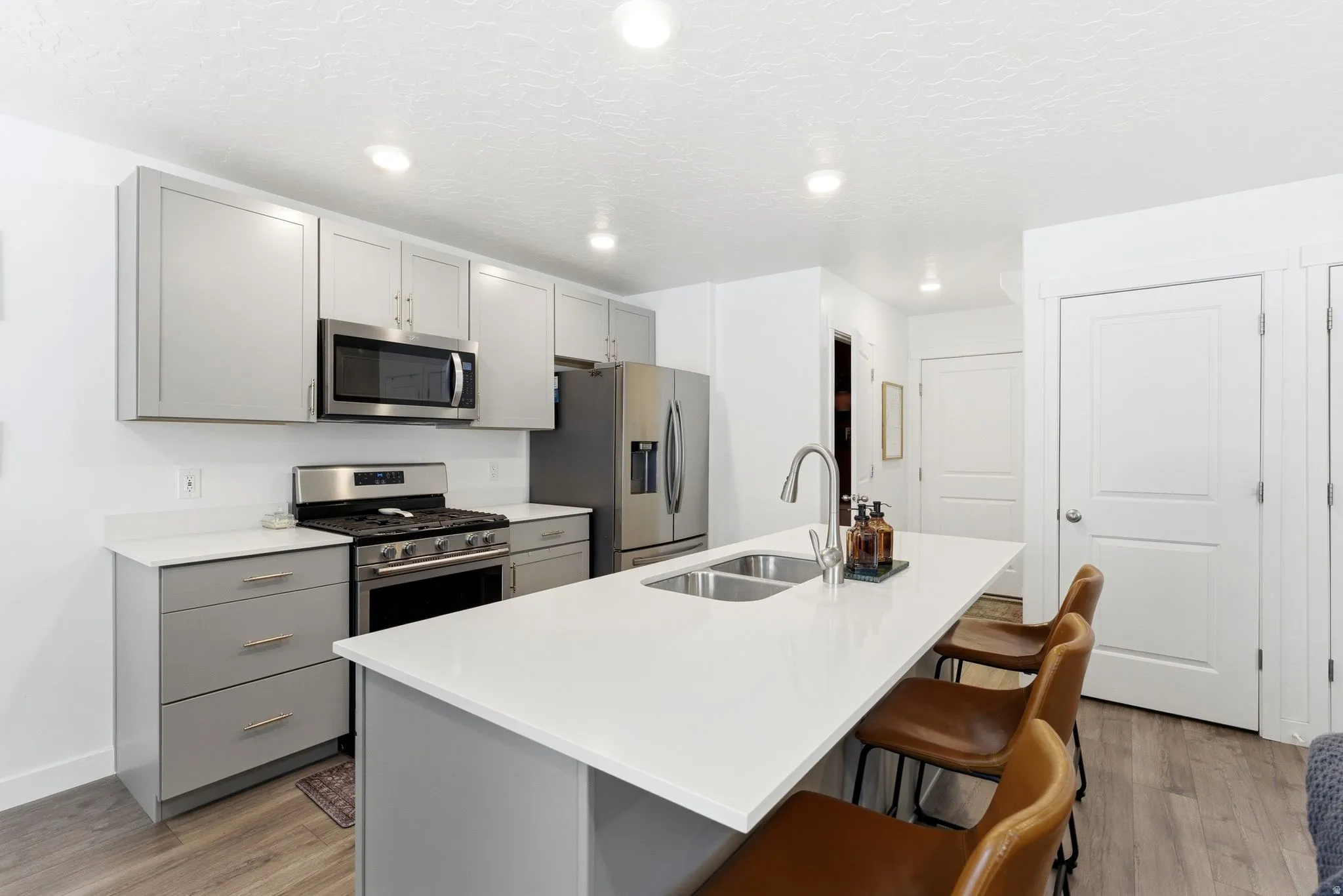 Modern kitchen featuring an island with a double sink, light gray cabinetry, stainless steel appliances, white countertops, and wood-finish flooring