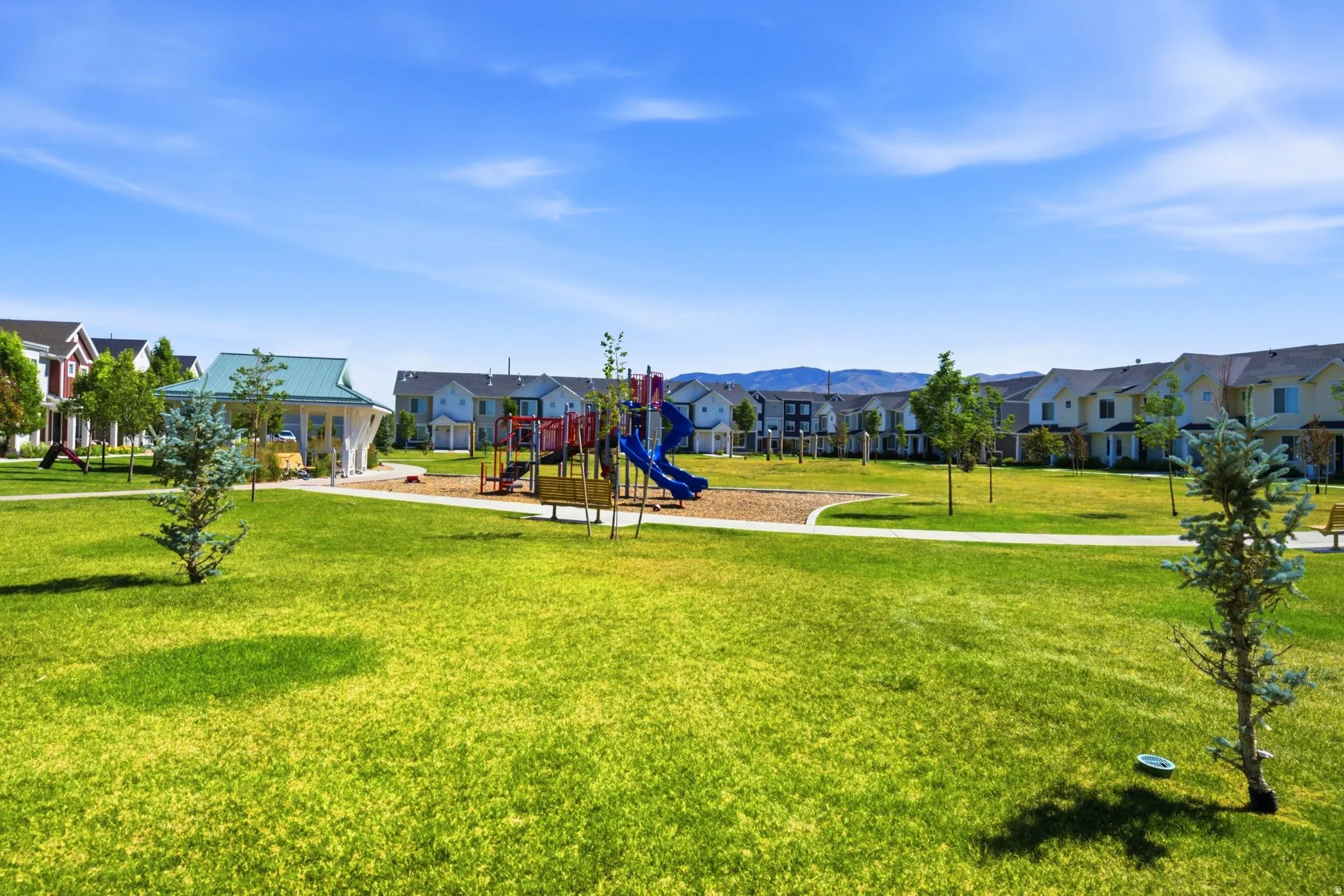 Expansive green space featuring a central playground, a covered gazebo, and a walking path, surrounded by multi-story residential structures and distant mountains