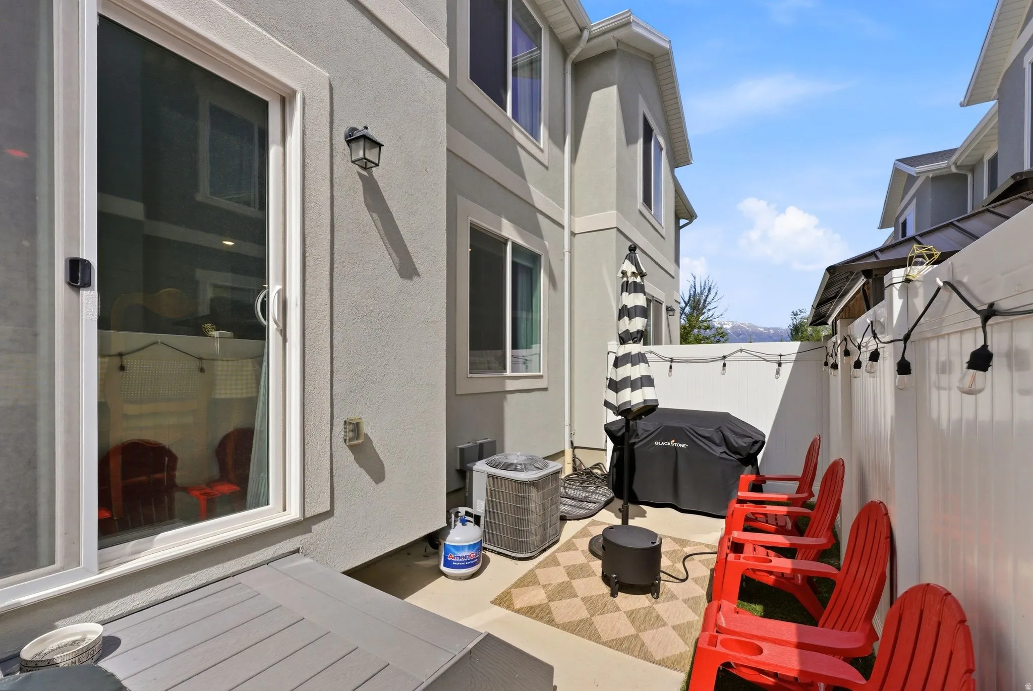 Private patio space with a neutral-toned exterior, featuring a sliding glass door, an exterior wall light fixture, and a white fence