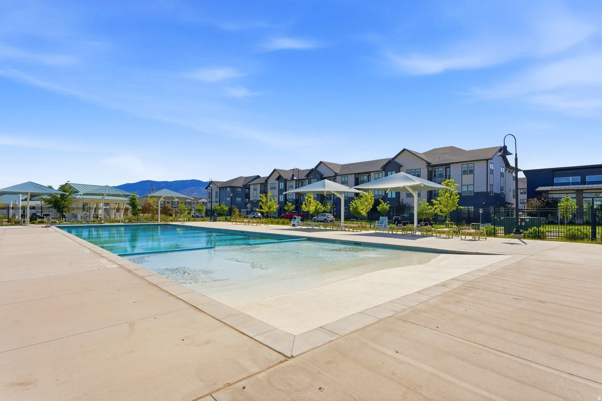 Community swimming pool with a shallow entry area, surrounded by a concrete deck and multiple shade structures