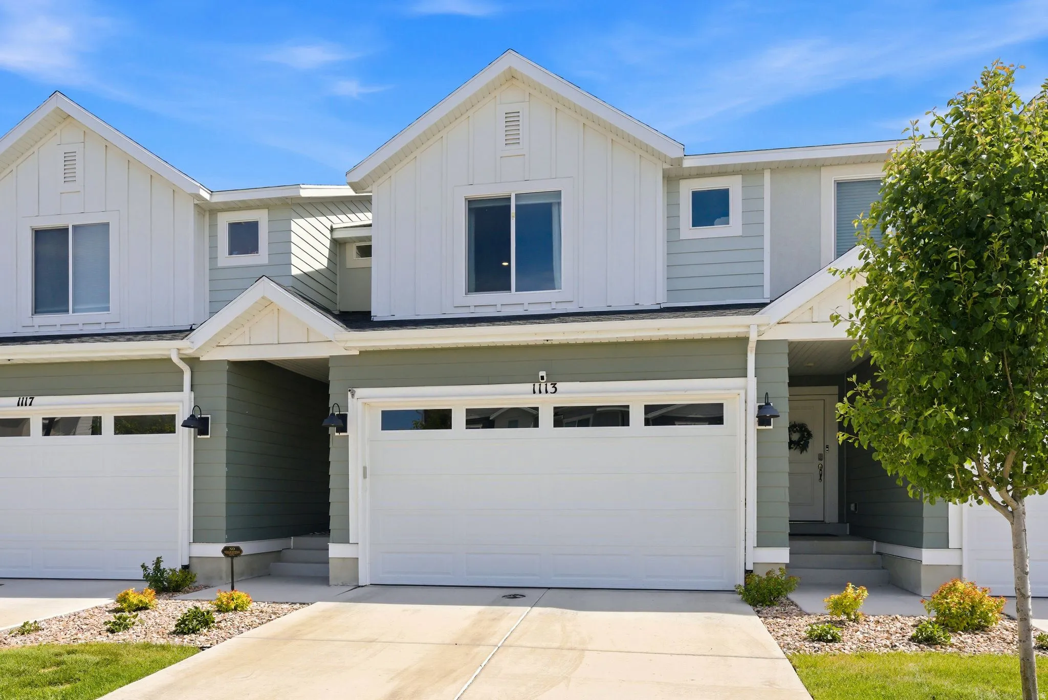 Modern townhouse exterior featuring a two-car garage, light green and white siding, a prominent gable roofline, and established landscaping