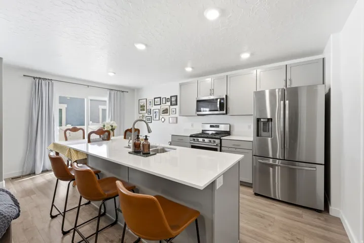 Open-concept kitchen featuring light wood-finish flooring, a central island with a sink and white countertop, light gray cabinetry, stainless steel appliances, and recessed lighting