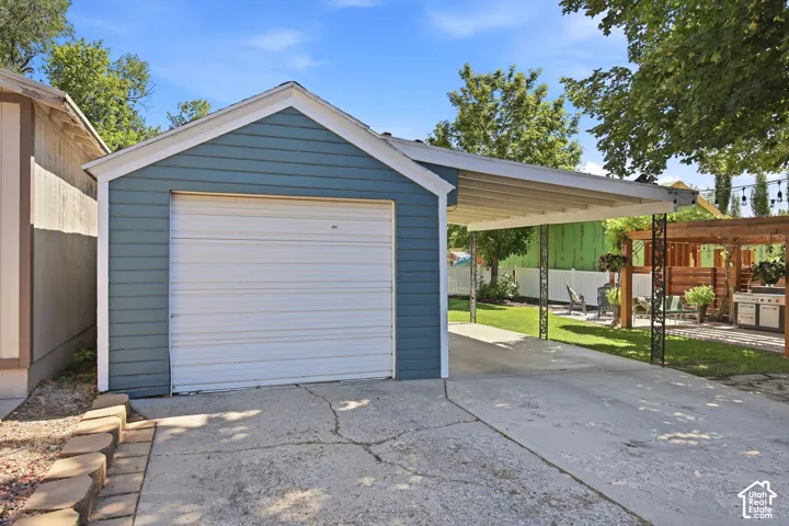 Detached garage featuring blue siding, a white garage door, and a concrete driveway leading to a carport with ornate metal supports