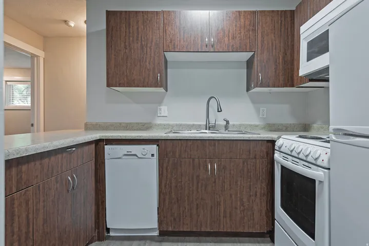 Kitchen featuring wood-finish cabinetry, light-toned countertops, a stainless steel sink with a gooseneck faucet, a white dishwasher, and a white electric range with an overhead microwave