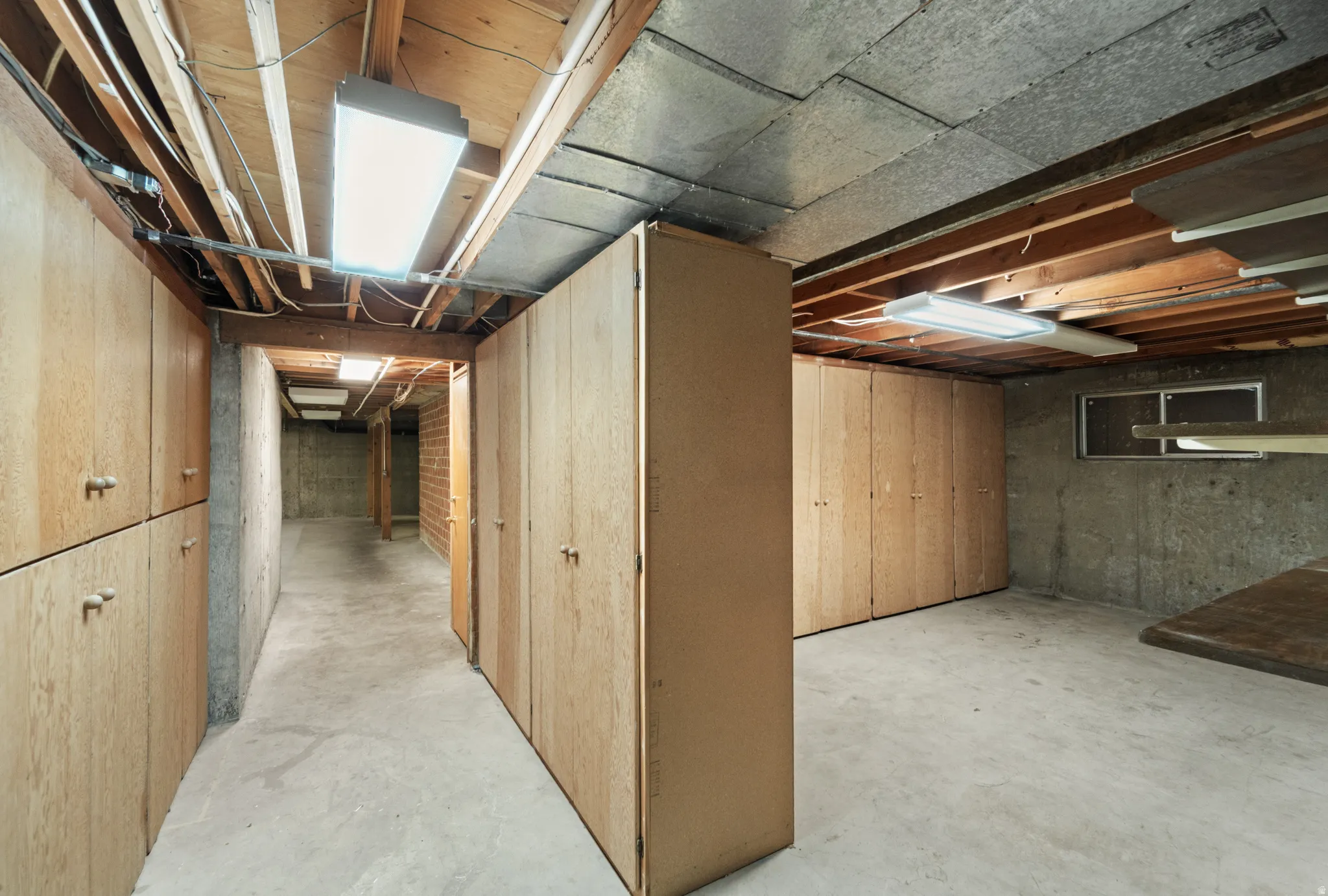 Basement storage area featuring extensive built-in cabinetry, exposed ceiling joists, concrete flooring, and fluorescent lighting