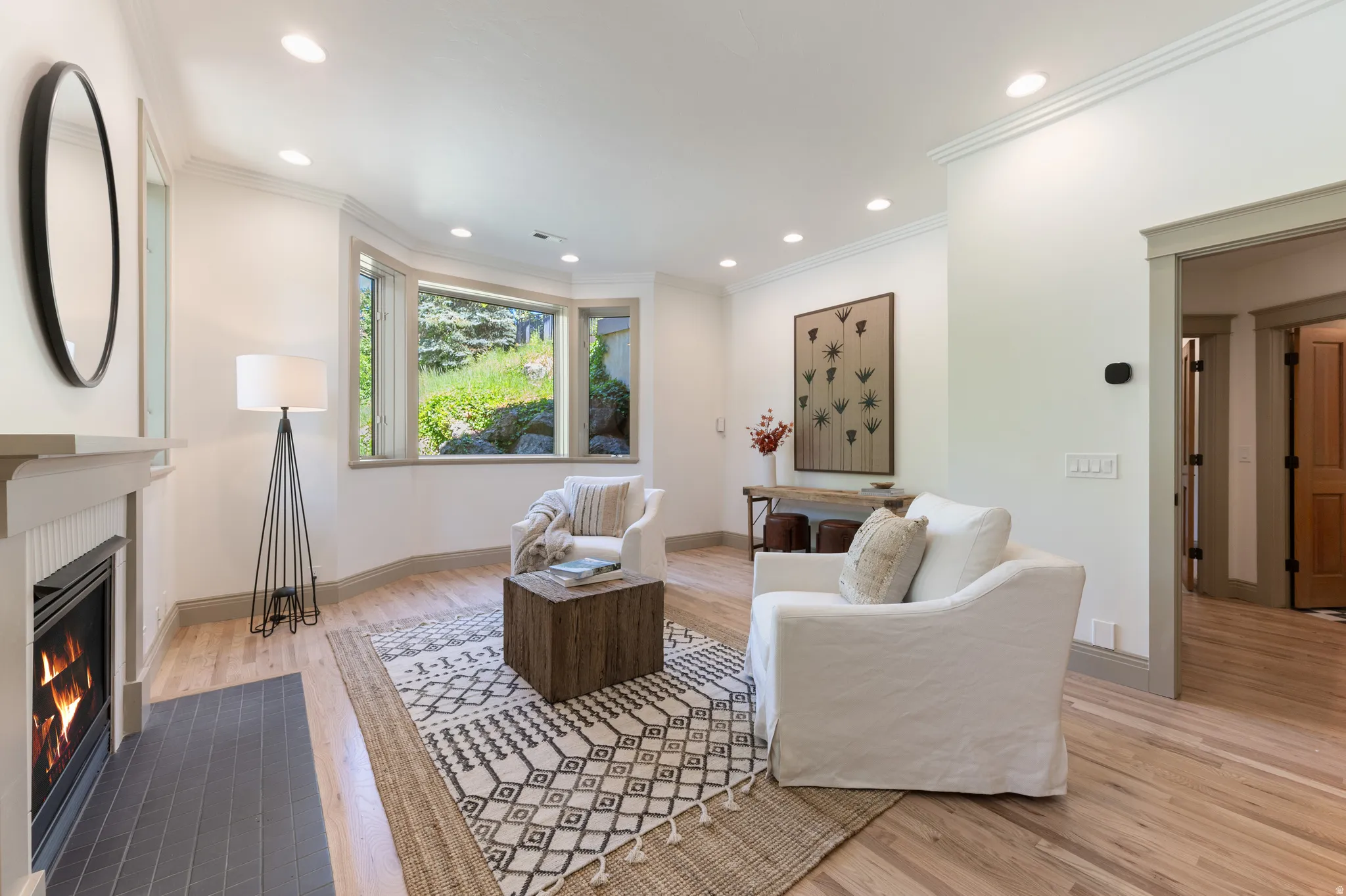 Recessed lighting illuminates this bright room featuring light wood-finish flooring, a bay window overlooking lush greenery, a modern fireplace with a tiled hearth, and crown molding