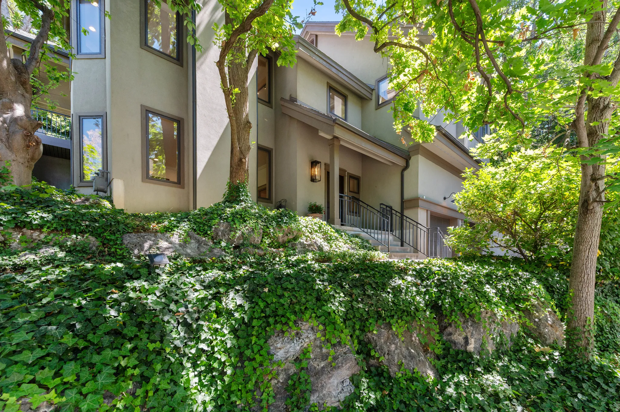 Stucco-sided residence with multiple levels, framed by mature trees and lush green ground cover
