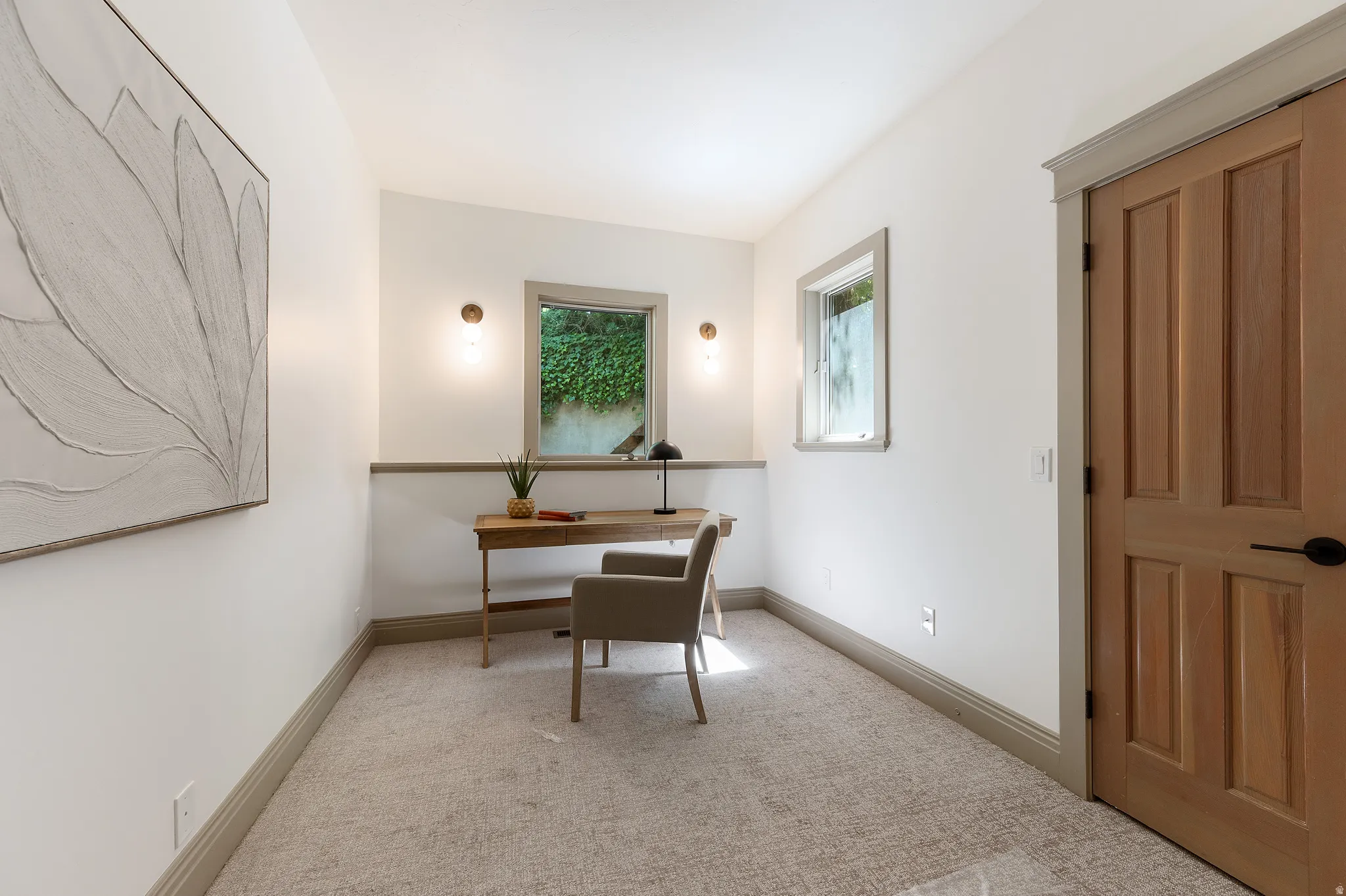 Carpeted room featuring a solid wood panel door with black hardware, two windows providing natural light, and two wall-mounted sconces