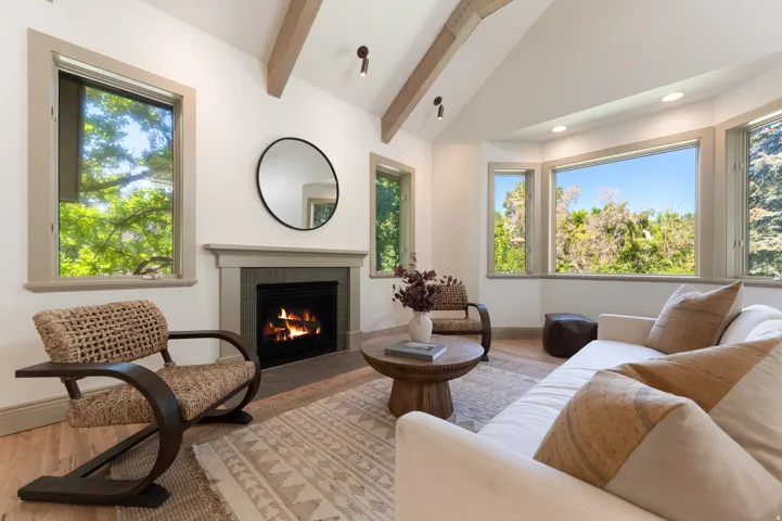 Vaulted ceiling featuring exposed wood beams, a fireplace with a stone mantel and tile surround, a bay window with a treetop panorama, and light wood-finish flooring