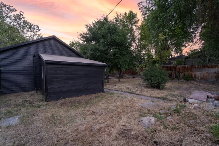 Exterior structure with black horizontal siding and an attached shed, situated on a lot with mature trees and a wood fence perimeter