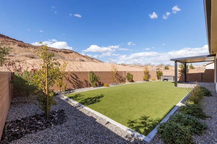 Expansive backyard featuring a manicured artificial lawn, a built-in patio cover, and desert landscaping with gravel and mature shrubs