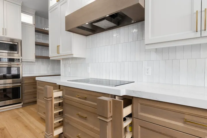 Integrated cooktop, stainless steel double ovens, white vertical tile backsplash, white countertops, and custom cabinetry with pull-out spice racks