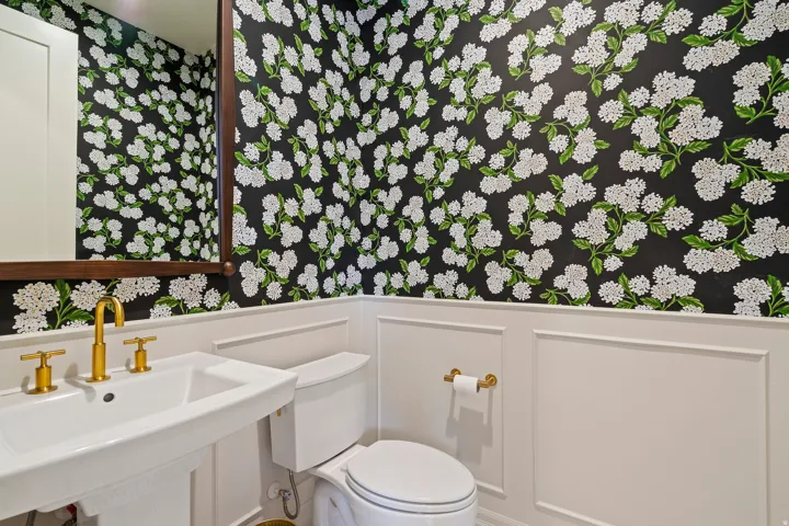Powder room featuring floral wallpaper, white wainscoting, a pedestal sink with brass fixtures, and a framed mirror