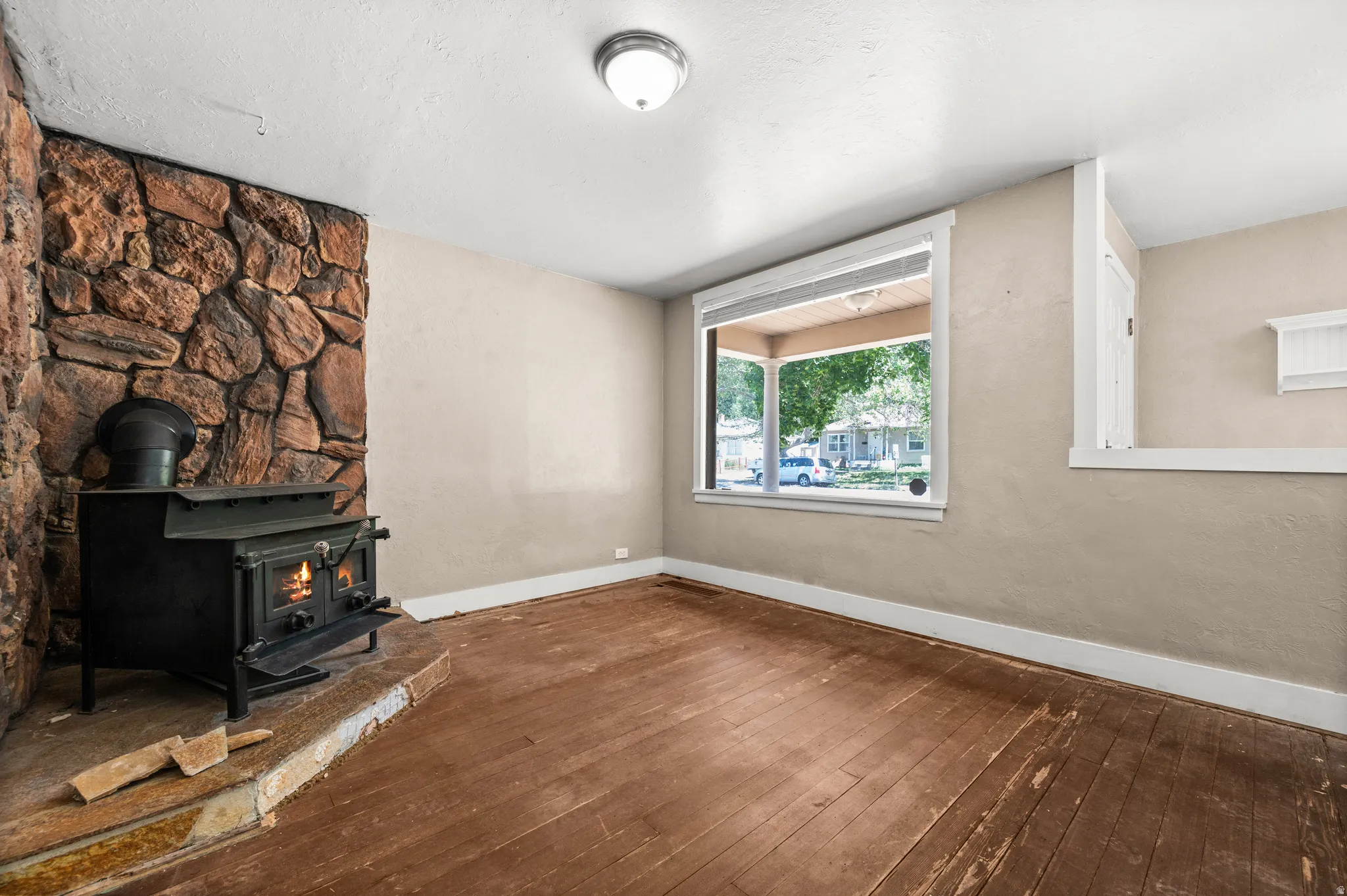Stone-faced fireplace with a black wood-burning stove, large picture window, and wood-finish flooring