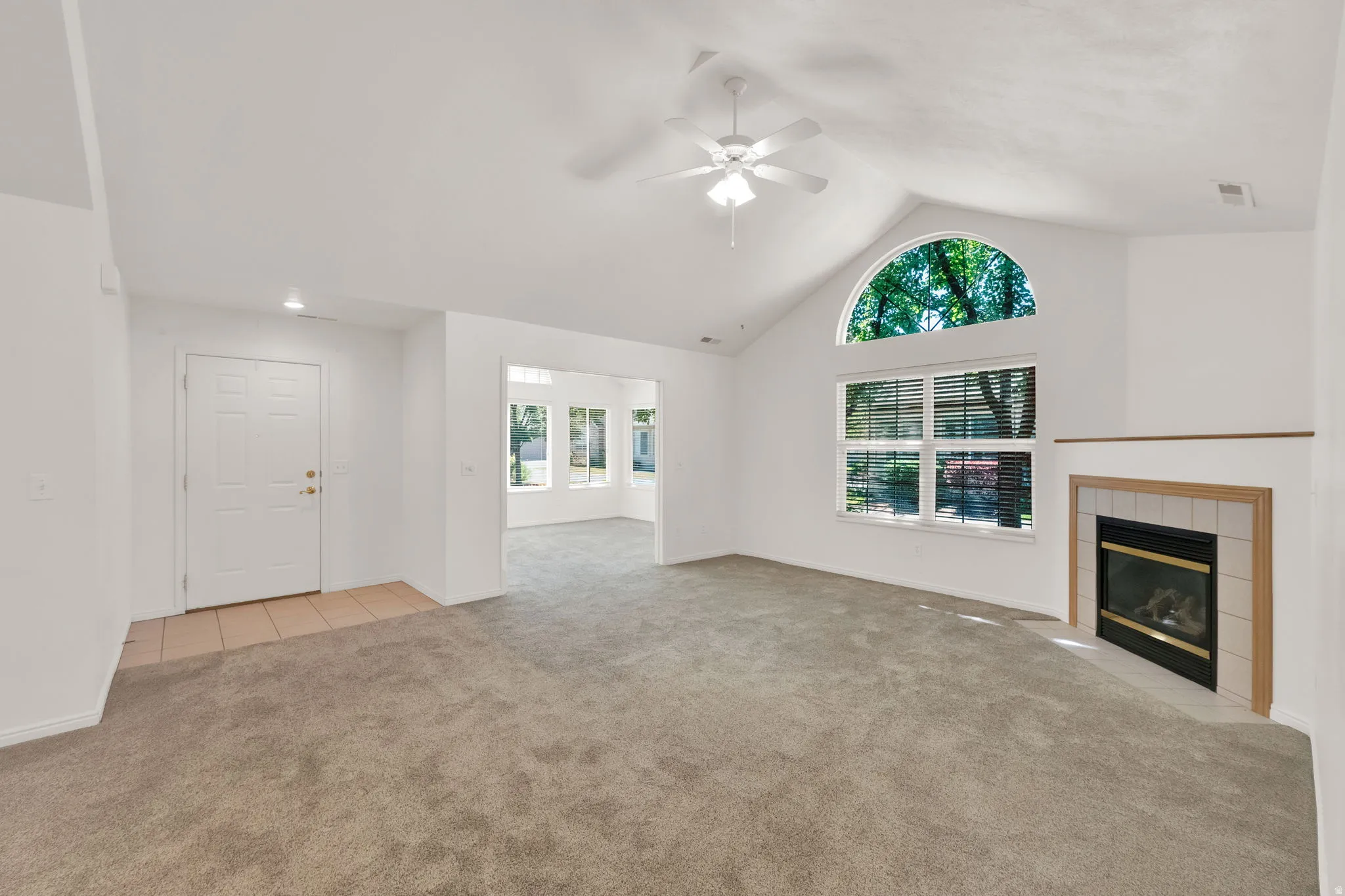 Spacious living area featuring vaulted ceilings with a ceiling fan, plush grey carpeting, and a fireplace with a tile surround and wood mantel
