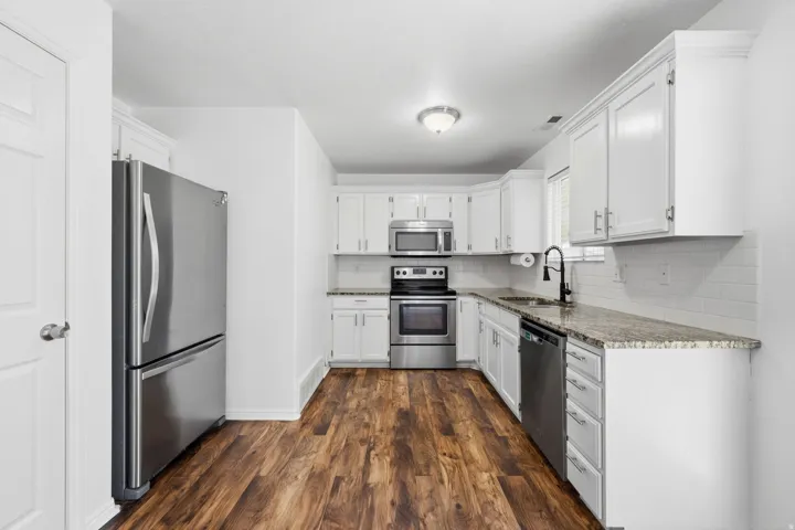 Modern kitchen featuring white cabinetry, granite-style countertops, stainless steel appliances, wood-finish flooring, and a subway tile backsplash