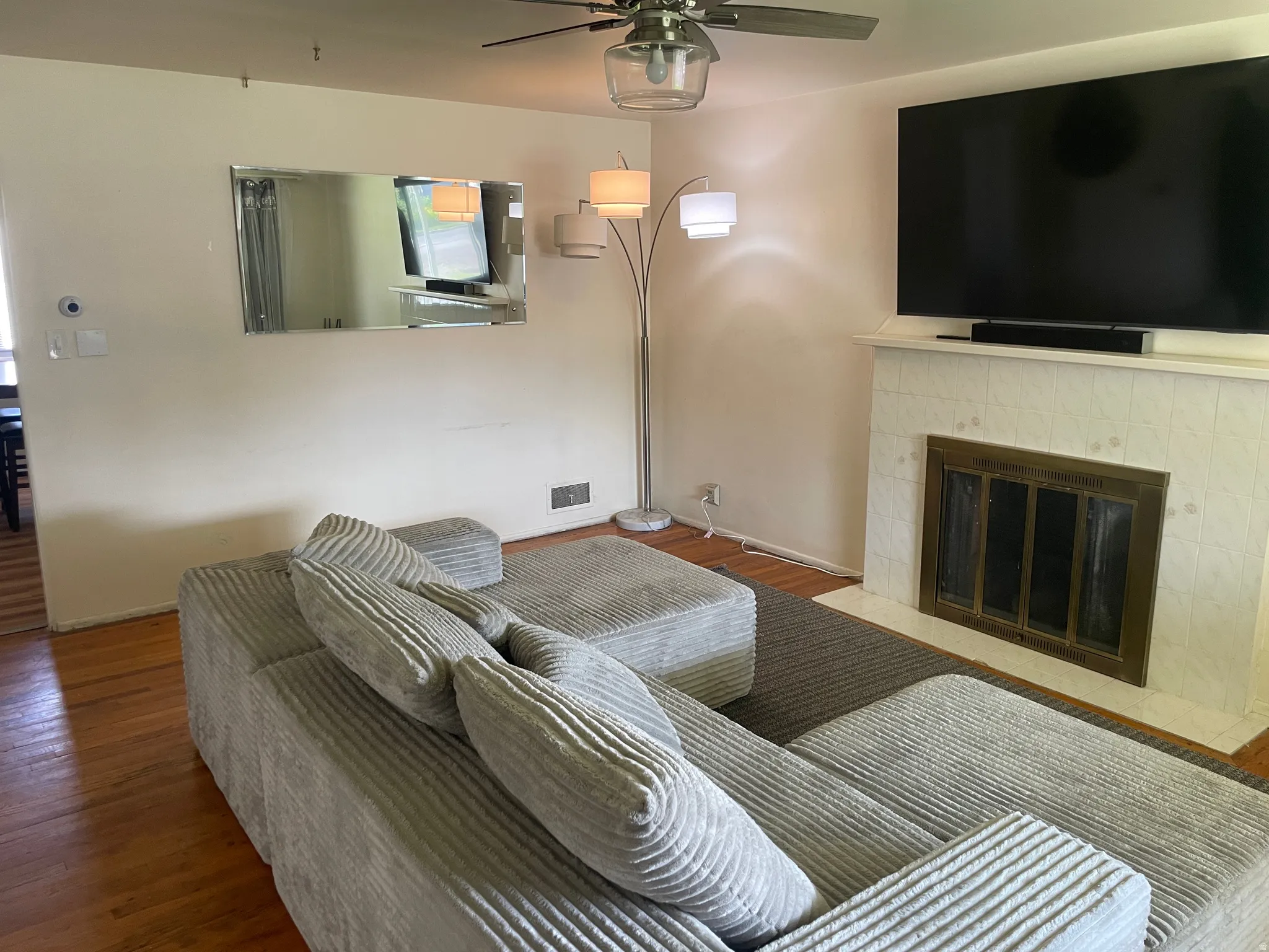 Living area featuring wood-finish flooring, a ceiling fan with integrated lighting, and a fireplace with a white tiled surround and brass-toned fire screen
