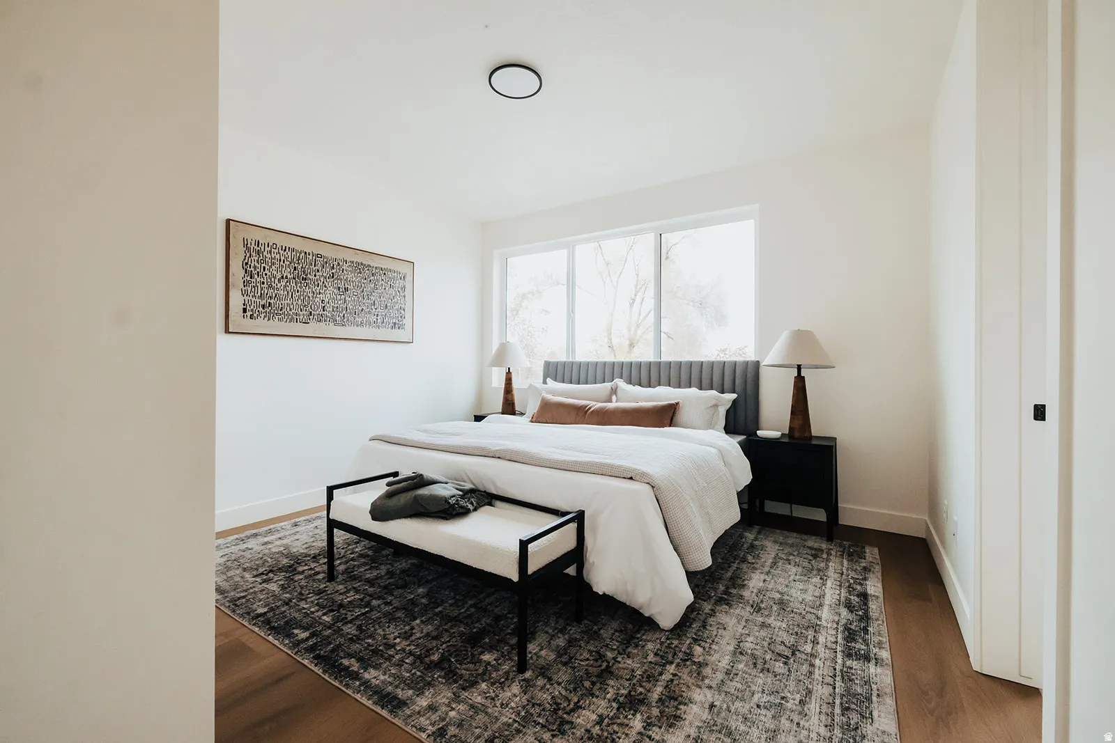 Spacious bedroom featuring light-toned walls, wood-finish flooring, a large horizontal window, and a flush-mount ceiling light fixture