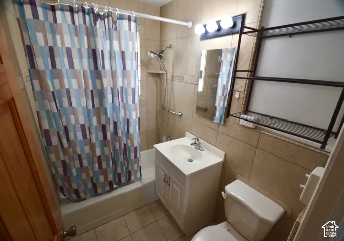 Bathroom featuring neutral-toned floor and wall tiling, a white vanity with integrated sink, a built-in bathtub with shower, and a wall-mounted mirror with vanity lighting