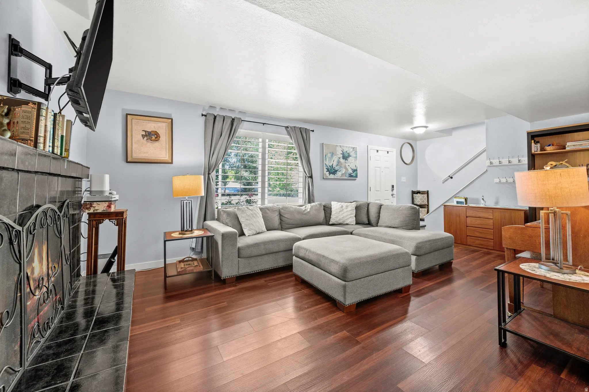 Spacious living area featuring rich wood-finish flooring, a tile-surround fireplace with an ornate screen, and a large window with plantation shutters