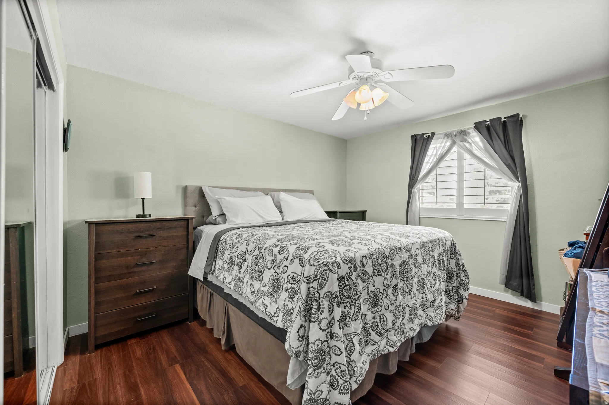 Bedroom featuring rich wood-finish flooring, a ceiling fan with integrated lighting, and a window with white plantation shutters