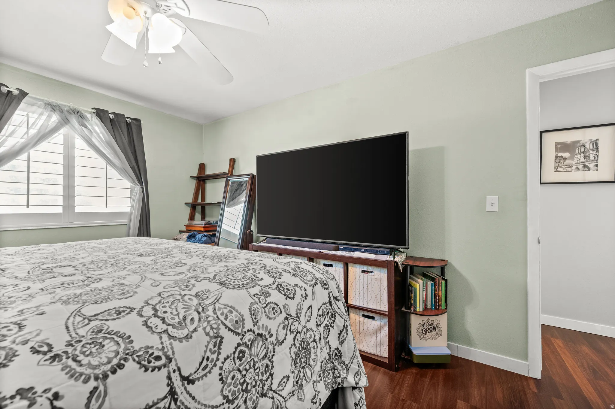 Bedroom featuring wood-finish flooring, a ceiling fan with integrated lighting, and a window with plantation shutters