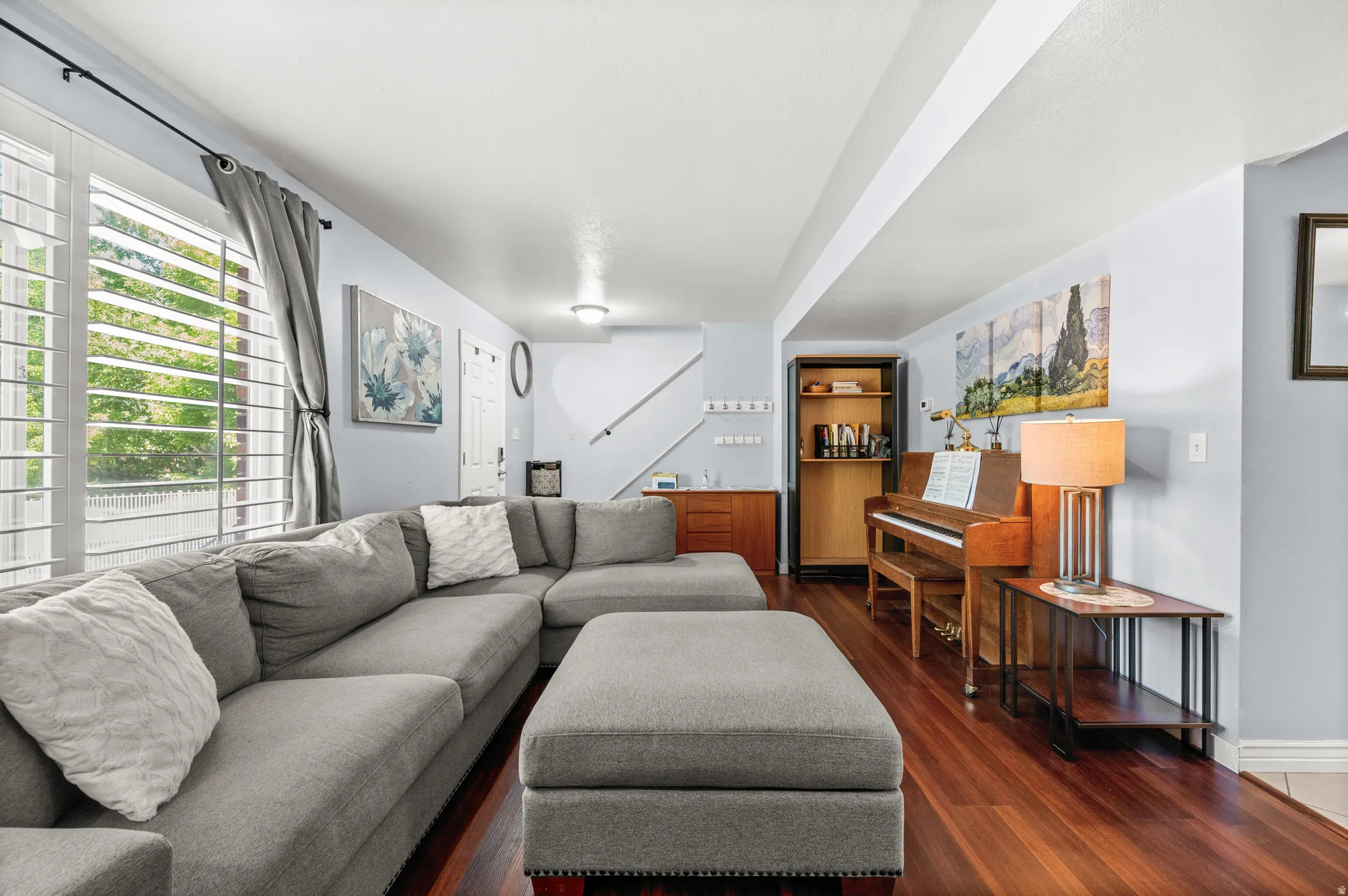 Spacious living area featuring rich wood-finish flooring, a large window with plantation shutters, light blue wall paint, and white trim