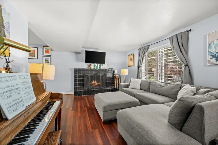 Living area featuring wood-finish flooring, a fireplace with a dark tile surround, a large window with plantation shutters, and light-toned walls