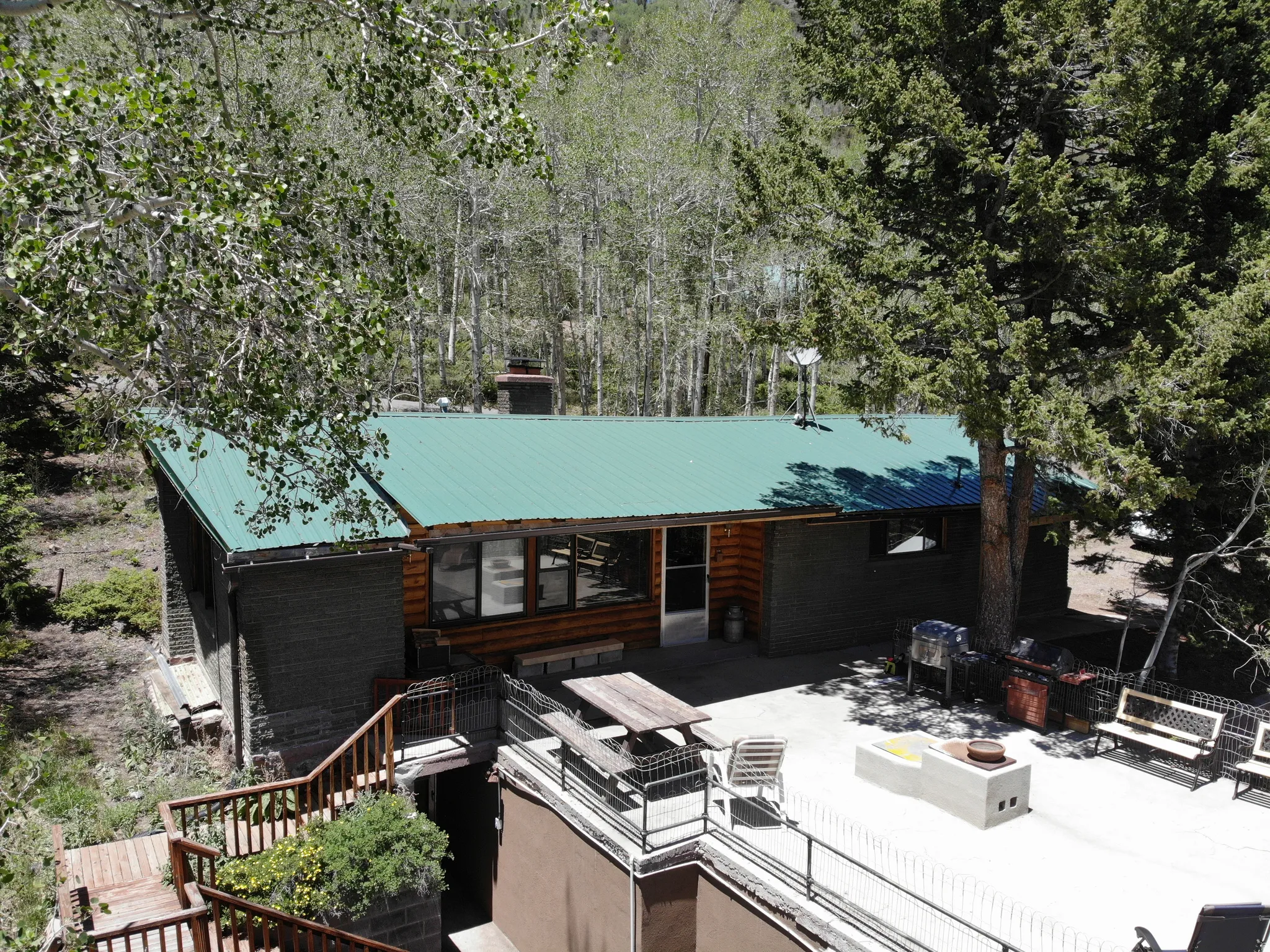 Log cabin exterior with a green metal roof, featuring a large concrete patio, a built-in outdoor fire pit, and a multi-level wooden deck with railing