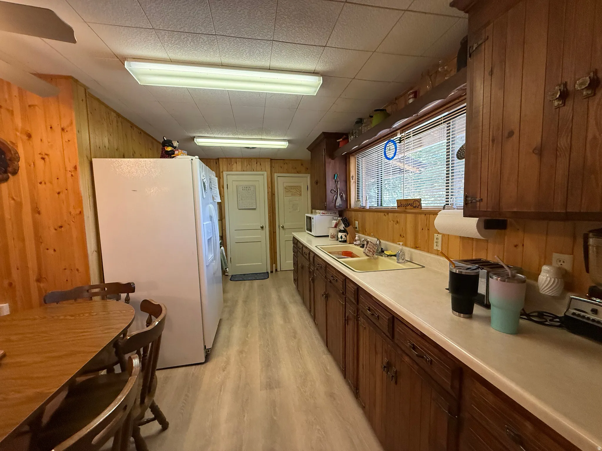 Kitchen featuring wood-finish flooring, wood paneling, a full-size window with blinds, upper and lower wood cabinetry, and a countertop with an integrated sink