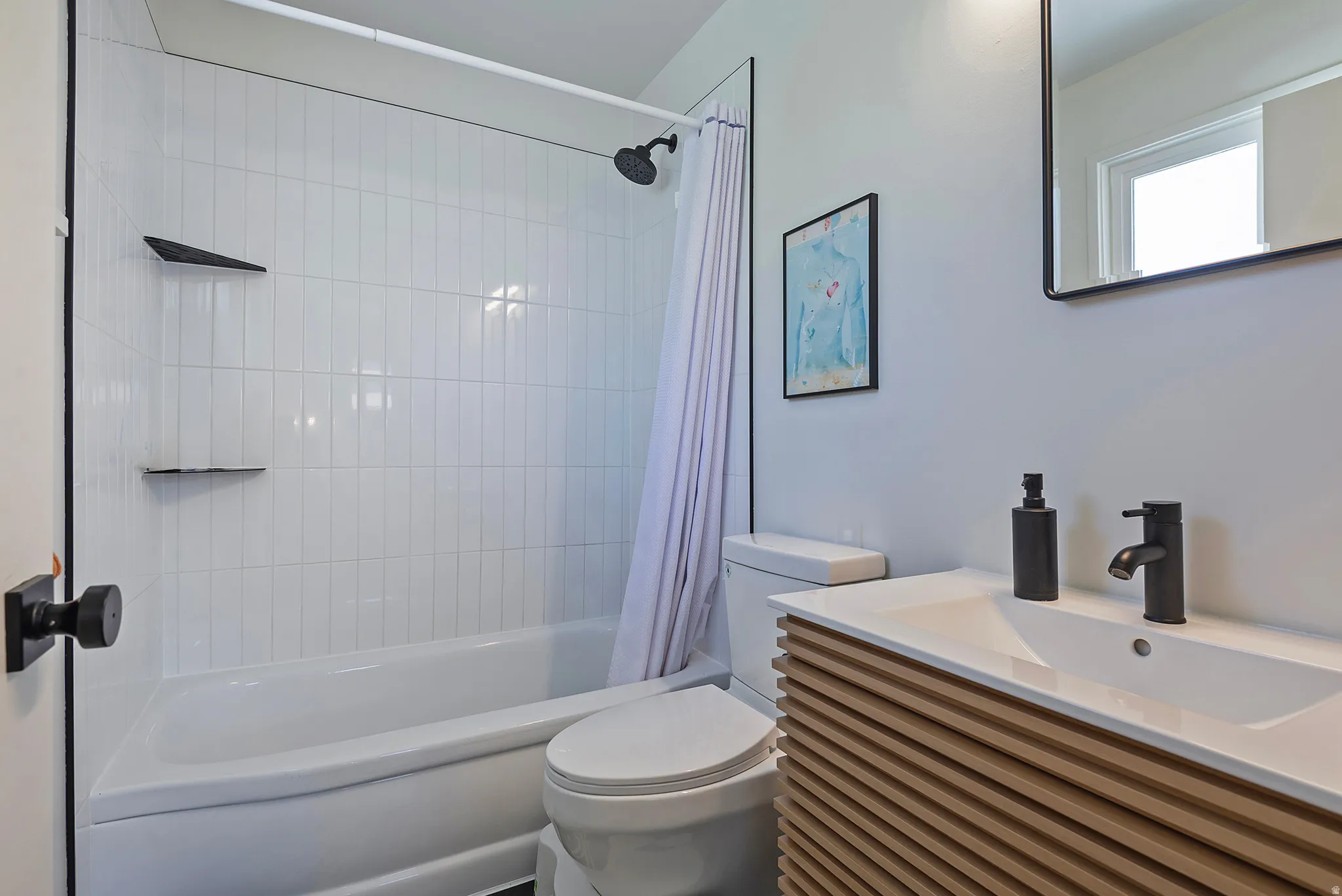 Bathroom featuring a white bathtub with vertical subway tile surround, matte black shower fixtures, a toilet, a light wood-finish vanity with an integrated white sink, and a matte black faucet