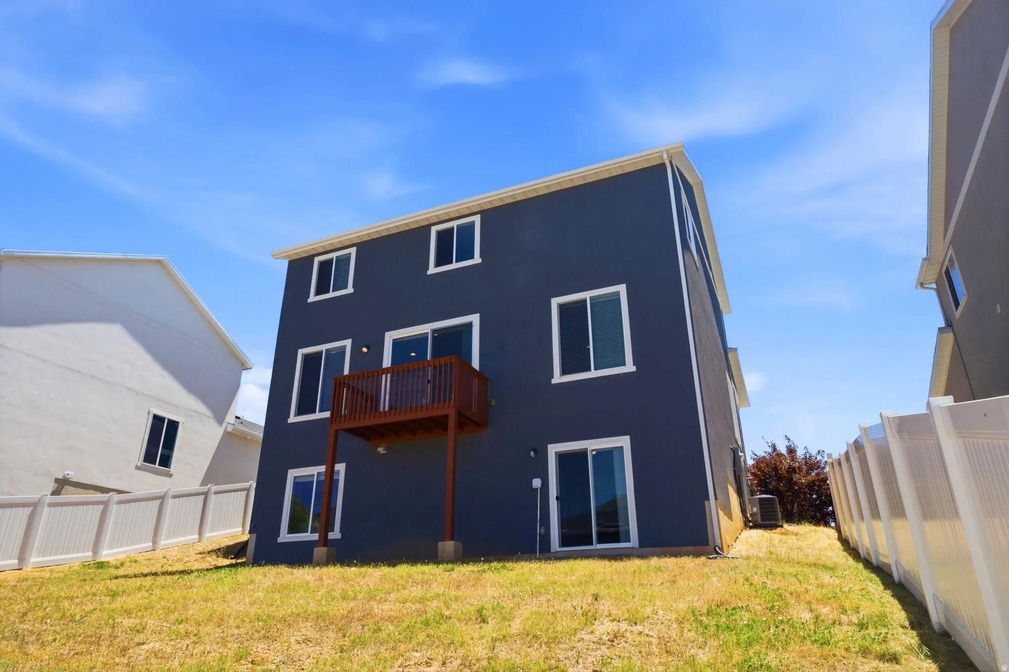 Rear exterior featuring a dark blue facade, multiple large windows, a second-story wooden deck, a walkout sliding glass door, and white vinyl fencing
