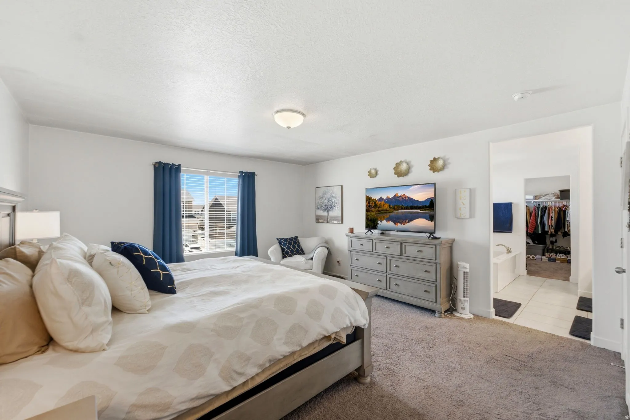 Carpeted room featuring a ceiling-mounted light fixture, window with blinds and drapes, and an open doorway revealing a tiled bathroom area with a built-in tub and closet