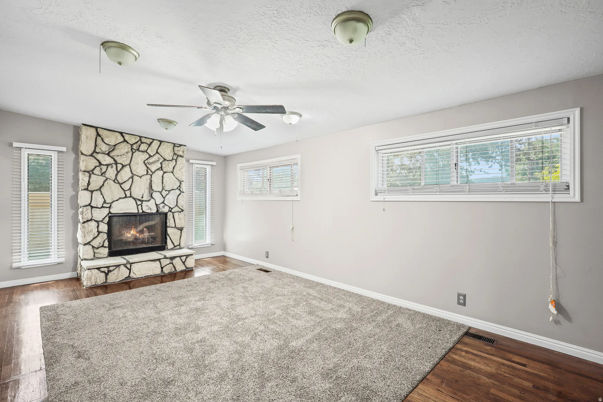 Living space featuring a stone-clad fireplace with a hearth, wood-finish flooring, and multiple windows with blinds