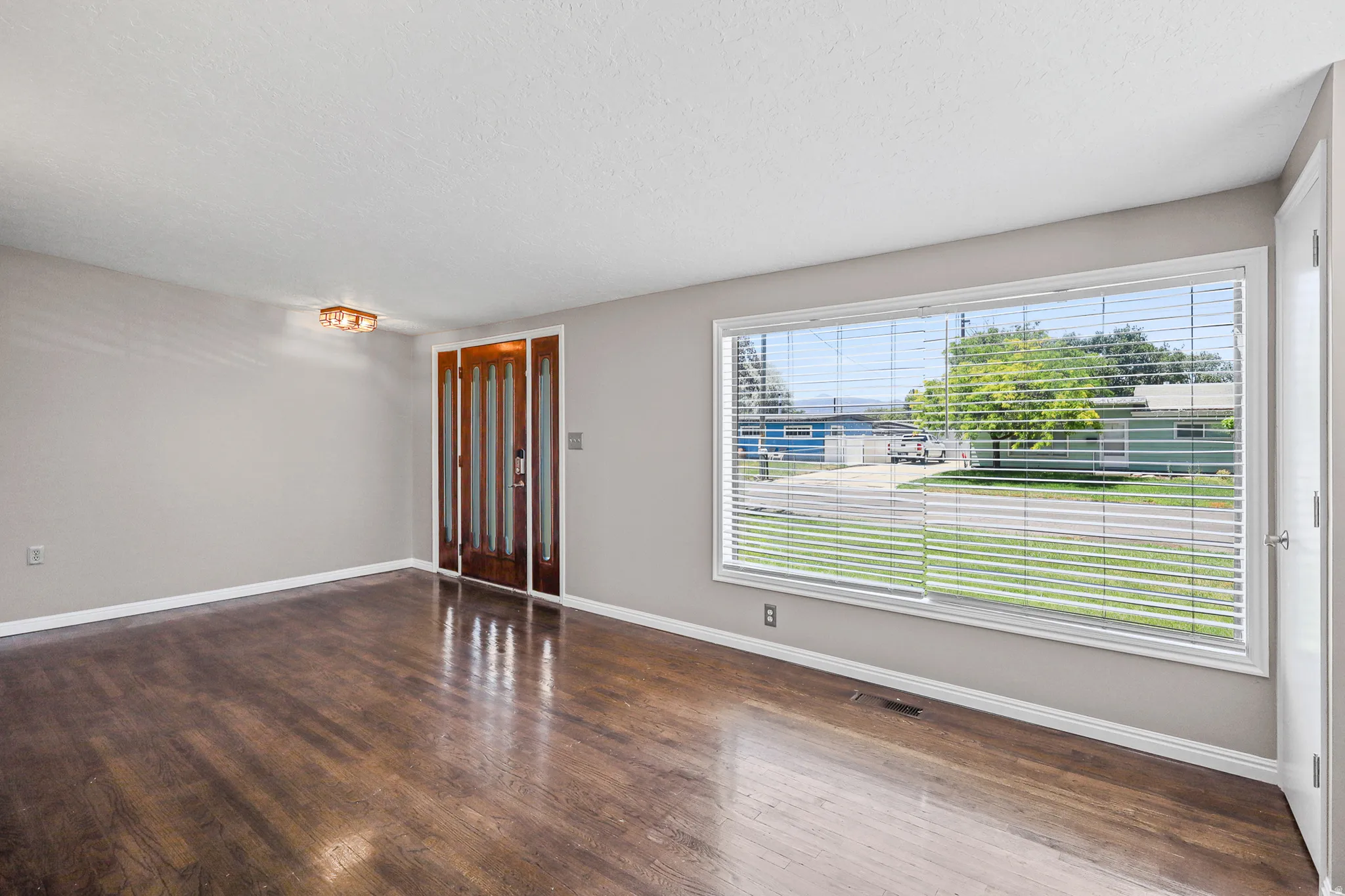 Spacious room featuring wood-finish flooring, a large picture window with horizontal blinds, and a paneled front door with sidelights